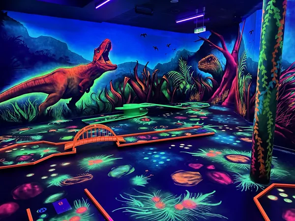 Schwarzlicht-Minigolfbahn mit großem T-Rex an der Wand und leuchtenden Urwaldpflanzen